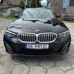 Przemysław Górski - Czarny samochód BMW, widok z przodu, z błyszczącą karoserią i tablicą rejestracyjną OK 9413C, zaparkowany na betonowym podłożu przed budynkiem.