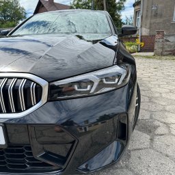 Przemysław Górski - Błyszczący, czarny samochód marki BMW, widok z bliska na reflektor i atrapę chłodnicy. Na karoserii odbija się otoczenie, w tle budynek z czerwonym akcentem.