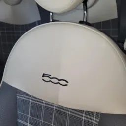 Detal wnętrza auta: białe sk&oacute;rzane siedzenie z logo '500', wykończone szarą kratą. Widoczny zagł&oacute;wek i fragment pas&oacute;w bezpieczeństwa. Jasne, r&oacute;wnomierne oświetlenie.