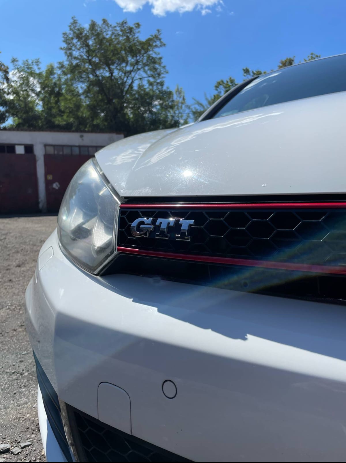 Biały samochód marki Volkswagen GTI z chromowanym logo na czarnej, heksagonalnej kratce z czerwoną obwódką, widok z dołu, na tle drzew i garażu.