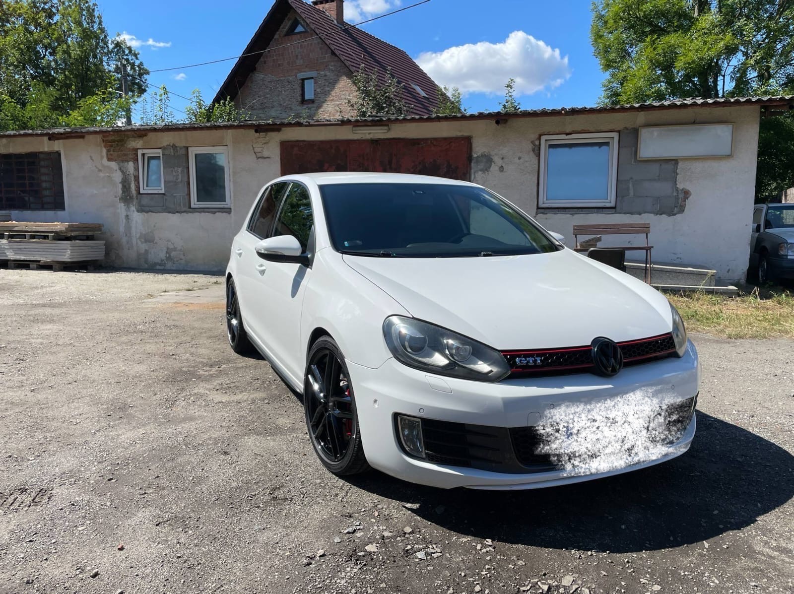 Biały Volkswagen Golf GTI, czarne felgi, czerwone akcenty. Auto stoi na żwirowym podłożu przed budynkiem z oknami. Jasne, słoneczne oświetlenie.