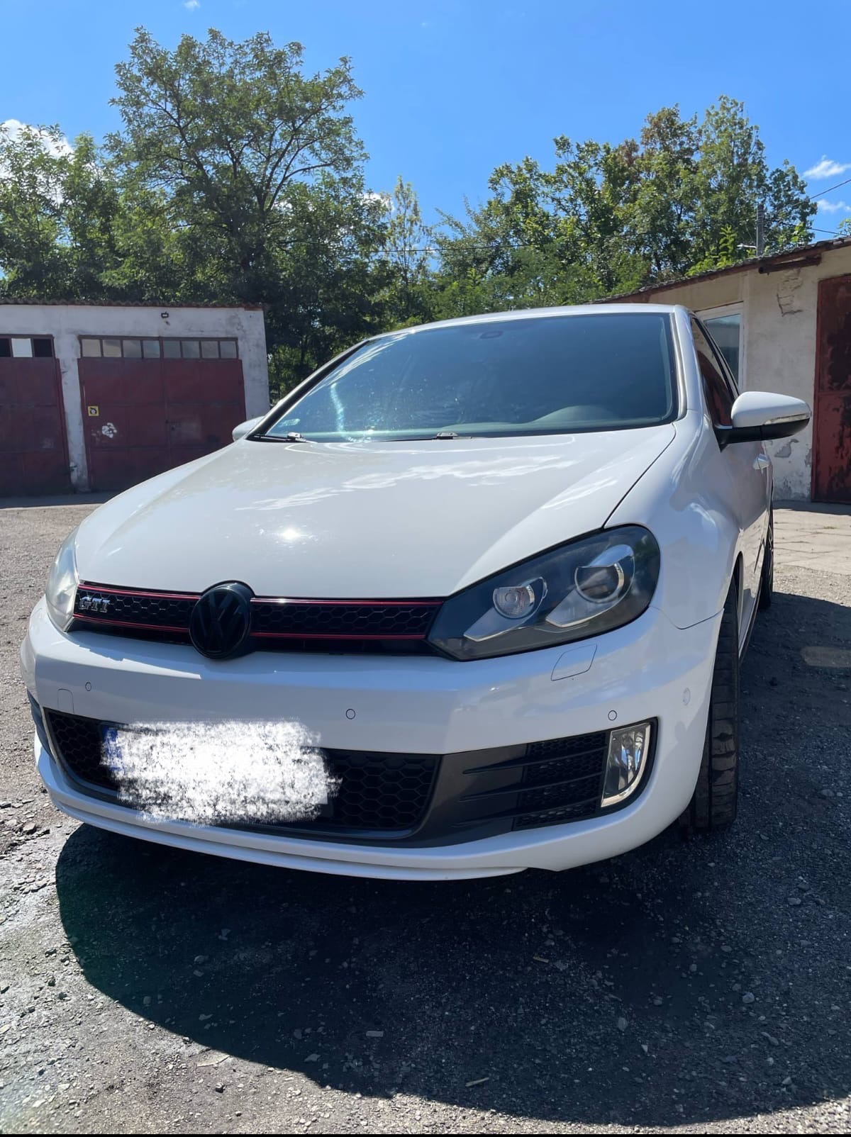 Biały Volkswagen Golf GTI, widok z przodu na zewnątrz, z zacienioną szybą i widocznymi detalami grilla. Tło garażu i zieleni.