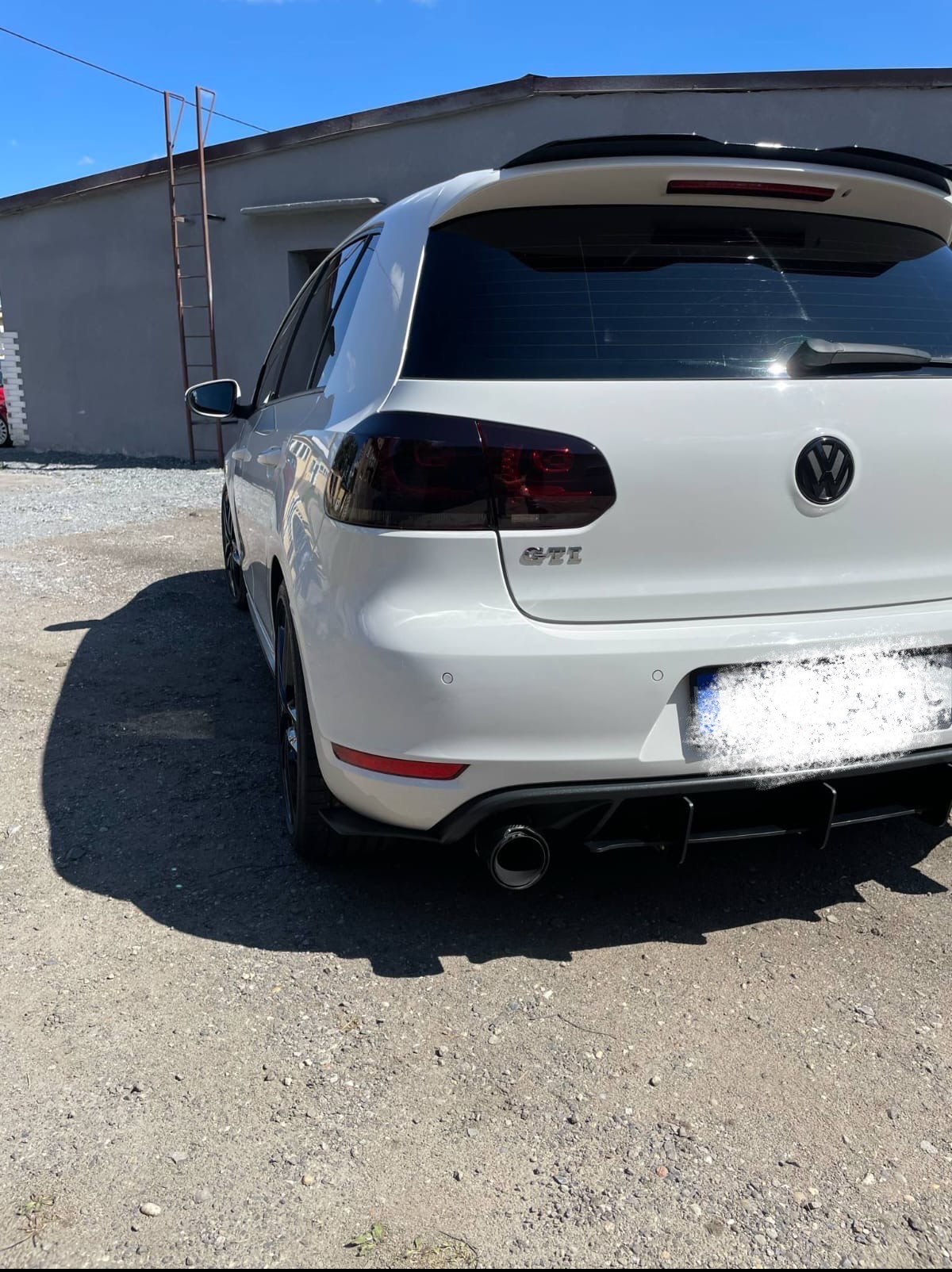 Biały Volkswagen Golf GTI z przyciemnionymi lampami, widziany z tyłu na żwirowym podłożu, w tle budynek z drabiną. Sportowy wygląd auta.