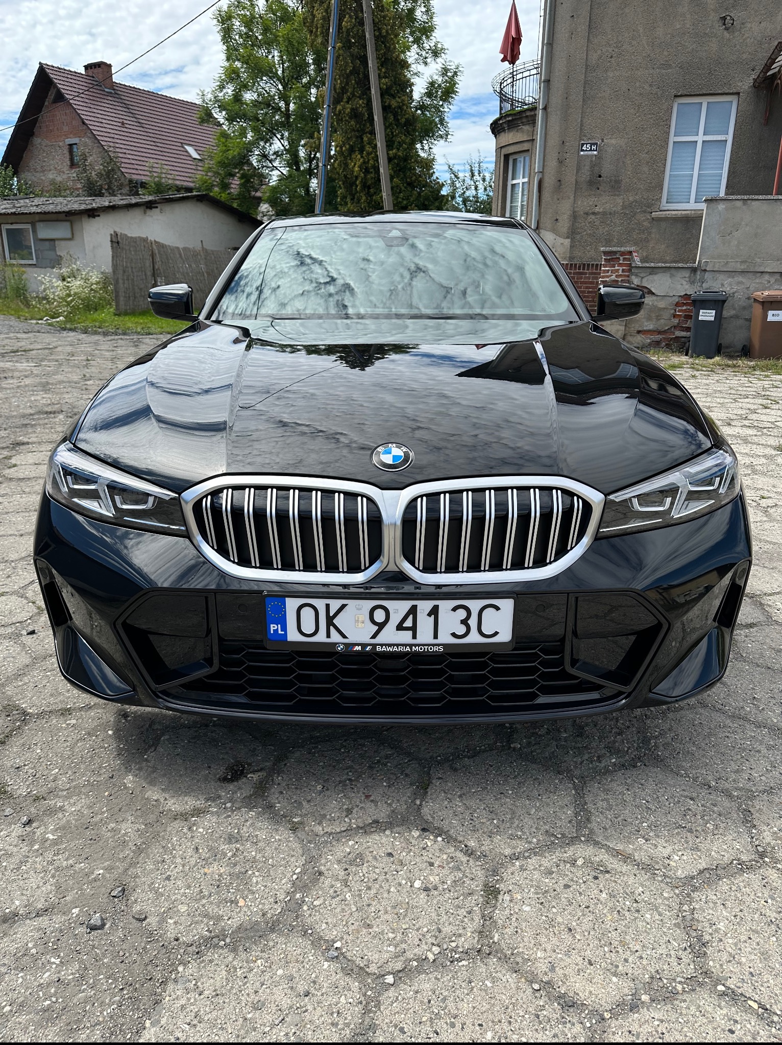 Czarny samochód BMW, widok z przodu, z błyszczącą karoserią i tablicą rejestracyjną OK 9413C, zaparkowany na betonowym podłożu przed budynkiem.