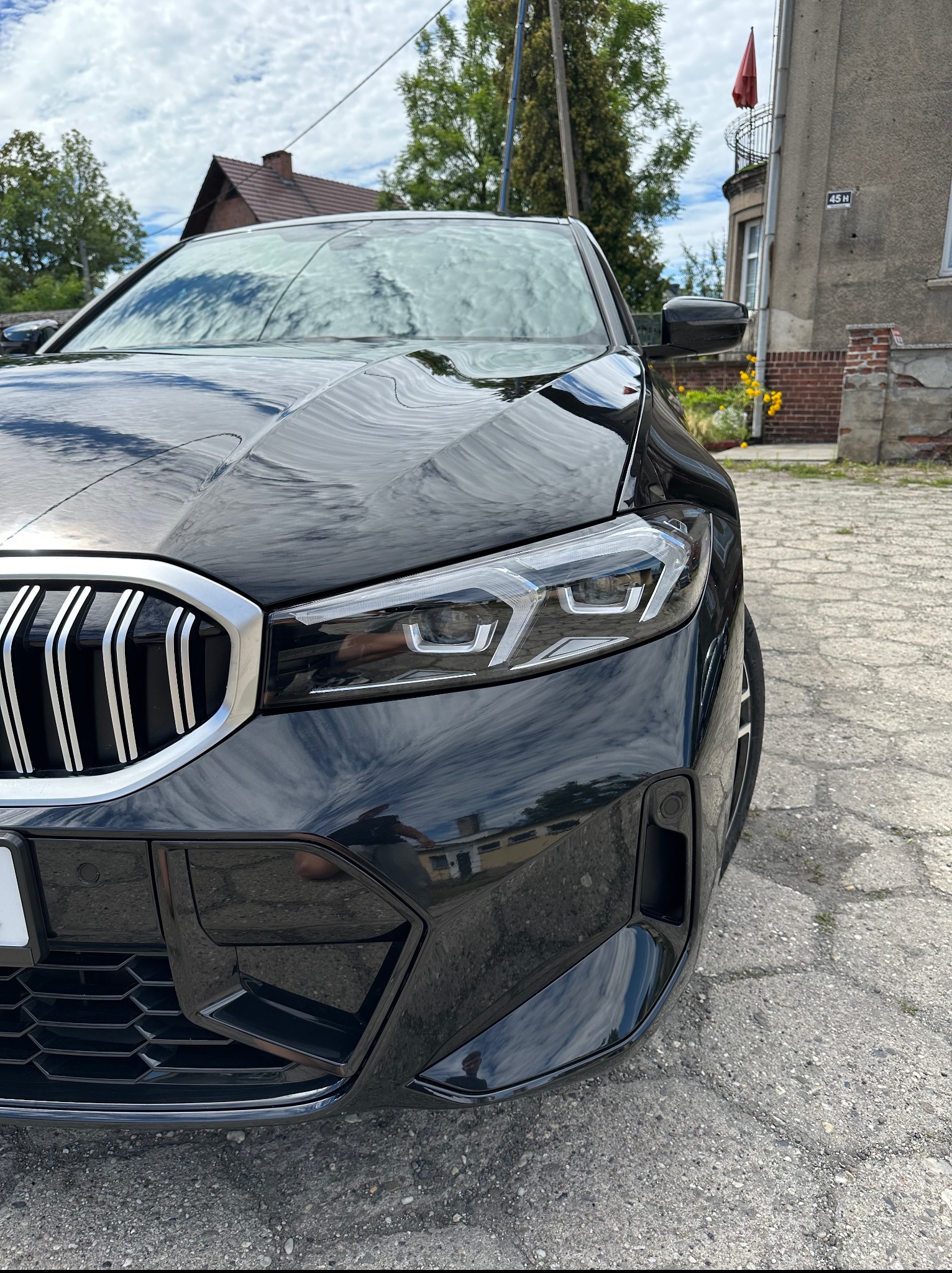 Błyszczący, czarny samochód marki BMW, widok z bliska na reflektor i atrapę chłodnicy. Na karoserii odbija się otoczenie, w tle budynek z czerwonym akcentem.