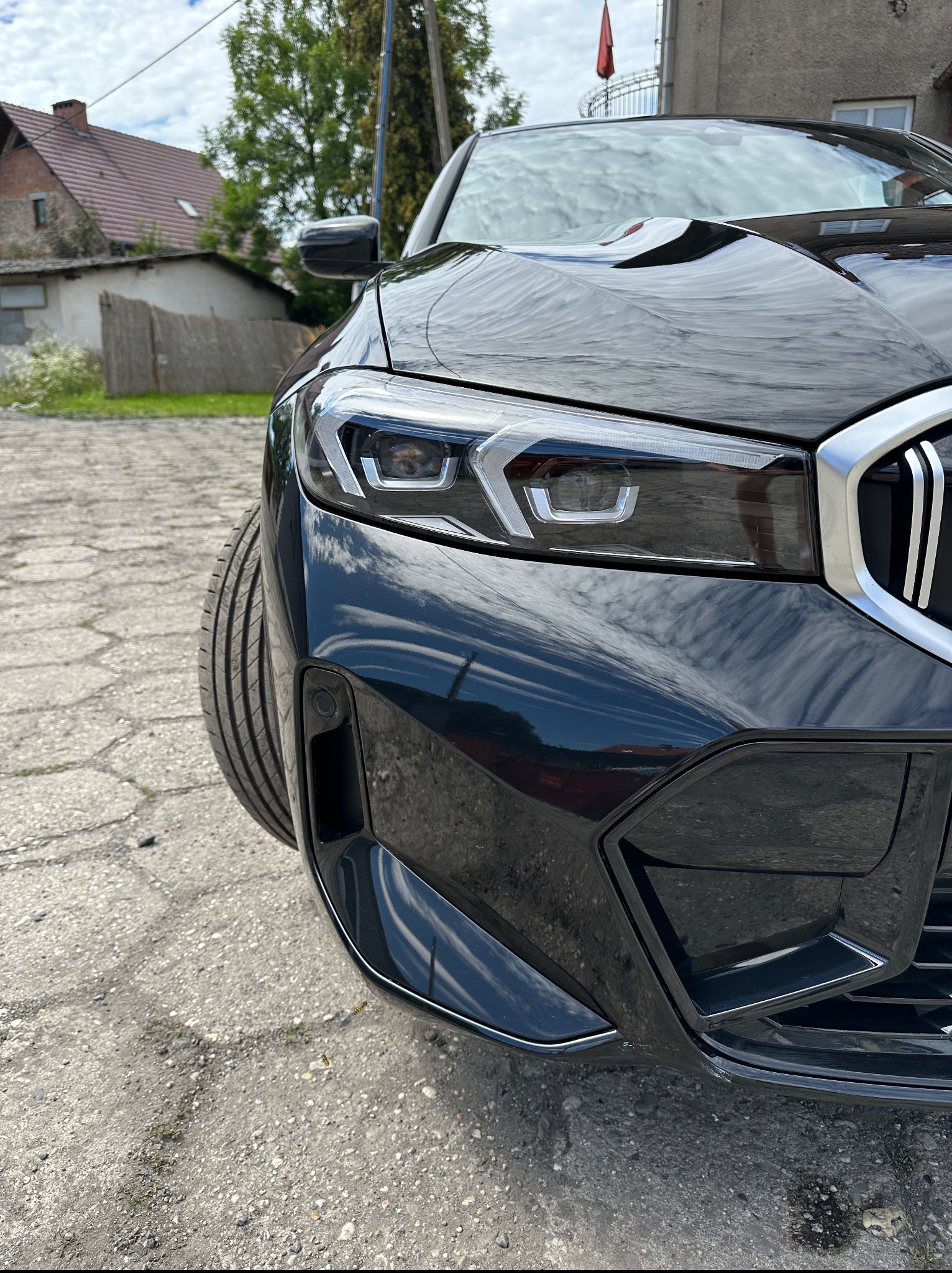 Błyszczący, czarny samochód marki BMW z nowoczesnym reflektorem, widoczny z bliska pod kątem, z odbiciem nieba w karoserii. Szara kostka brukowa w tle.