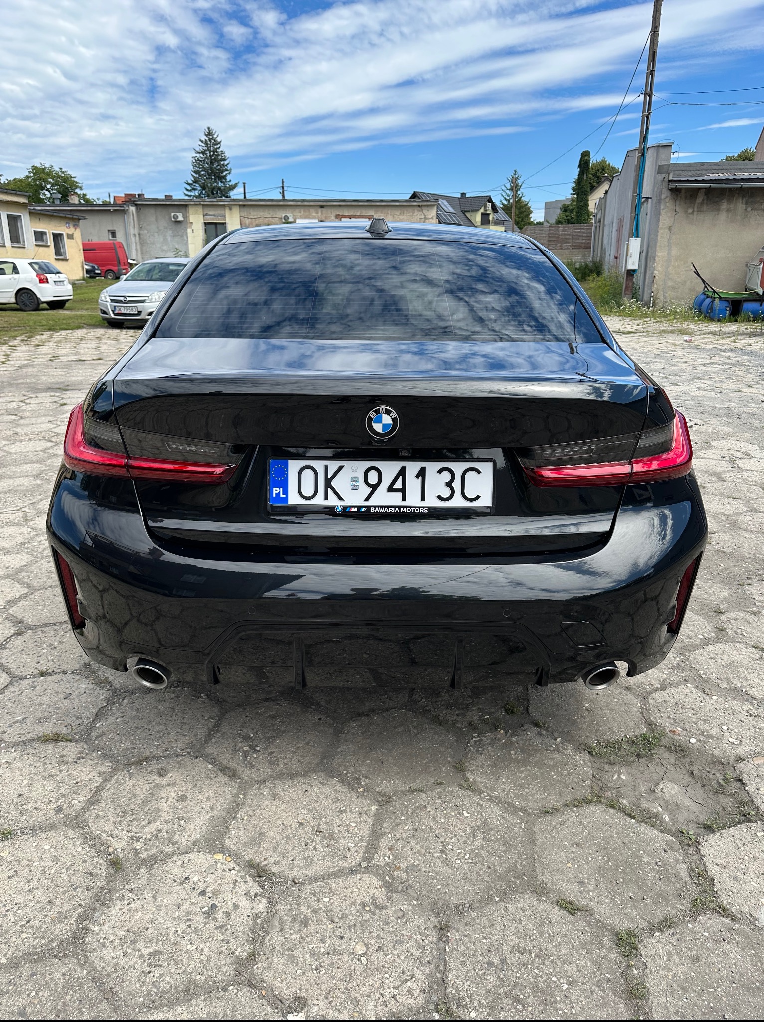 Czarne BMW serii 3, widok z tyłu na betonowym placu. Widoczne tylne światła, tablica rejestracyjna OK 9413C i logo BMW. W tle budynki i błękitne niebo.