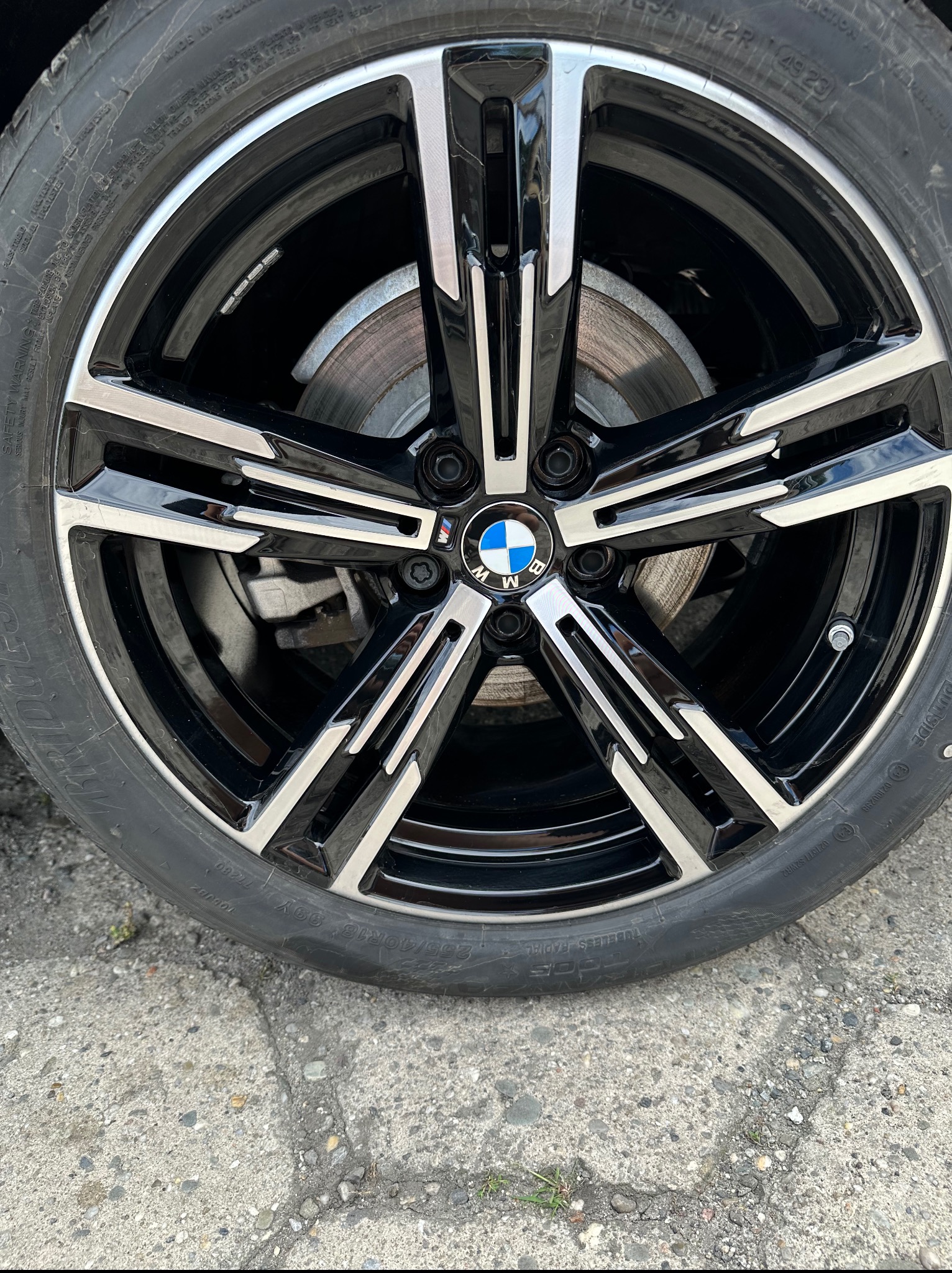 Błyszcząca, czarno-srebrna felga BMW z oponą na szarym, betonowym podłożu. Widoczne detale bieżnika opony i tarczy hamulcowej.