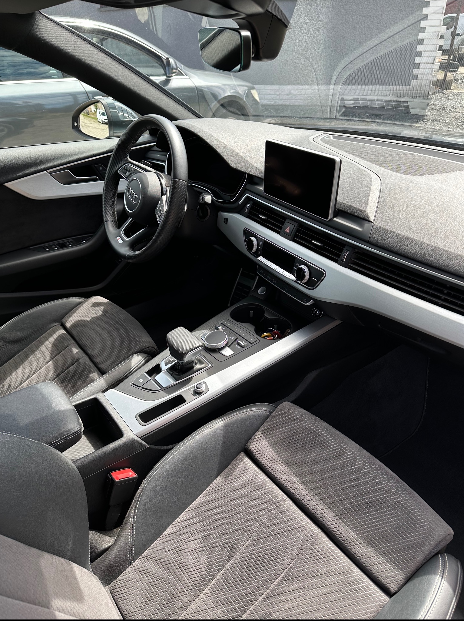 Wnętrze samochodu Audi z szarą tapicerką, widok z perspektywy pasażera, ukazujący deskę rozdzielczą, kierownicę i fotele. Jasne oświetlenie podkreśla detale wykończenia.