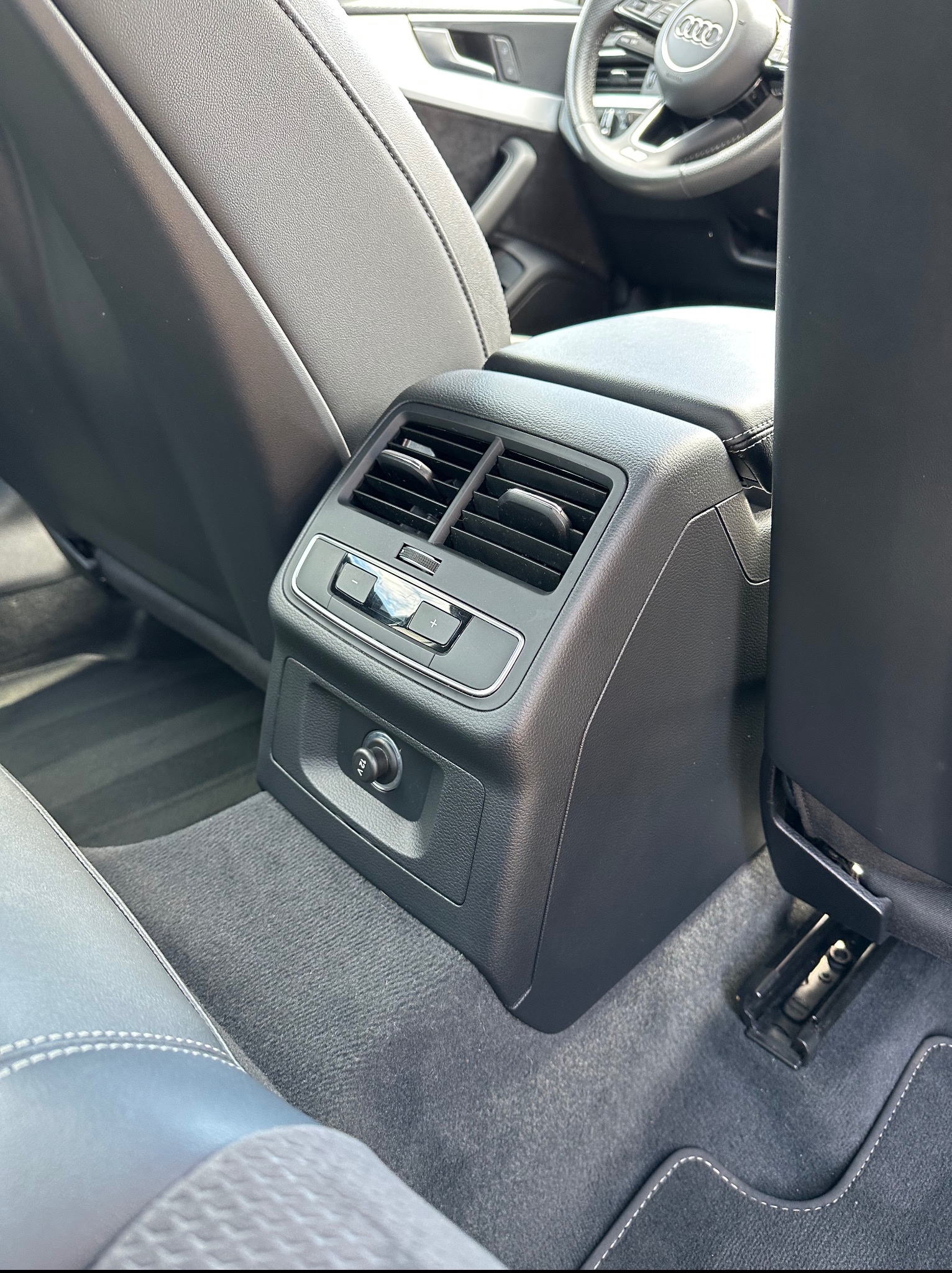 Wnętrze samochodu Audi: czarna skórzana tapicerka, tylny panel klimatyzacji z gniazdem 12V, szara wykładzina podłogowa z białym szwem. Widok na kierownicę z logo.