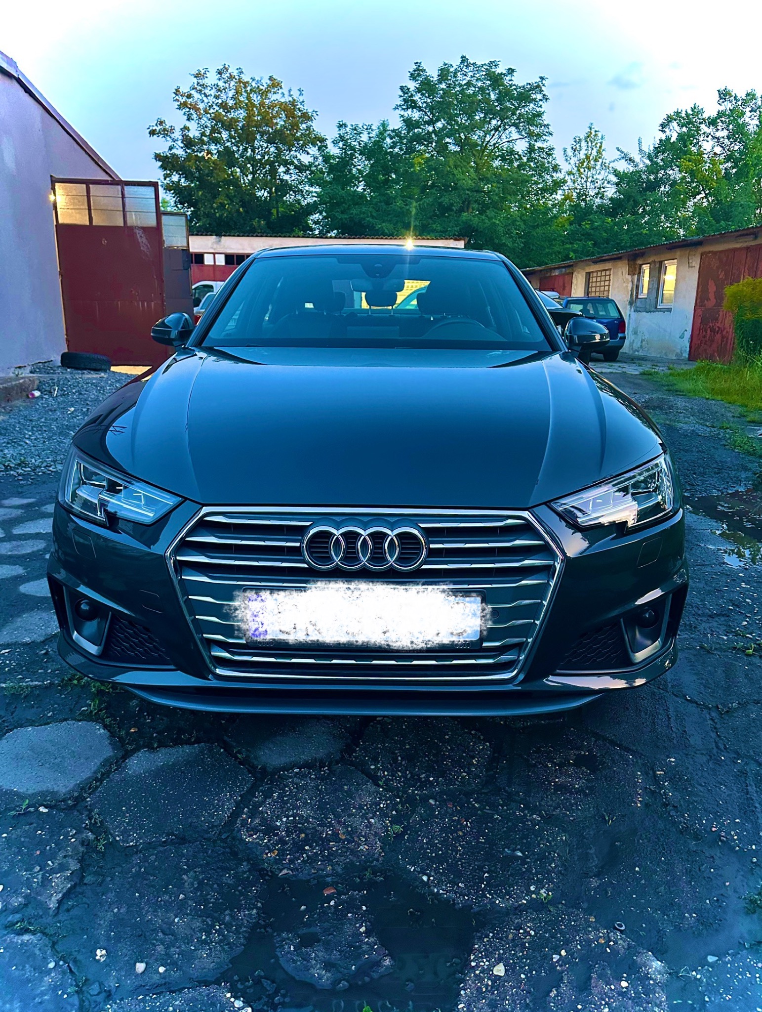 Ciemny samochód Audi A4, widok z przodu na mokrej kostce brukowej, z budynkami i zielenią w tle. Tablica rejestracyjna jest zamazana.
