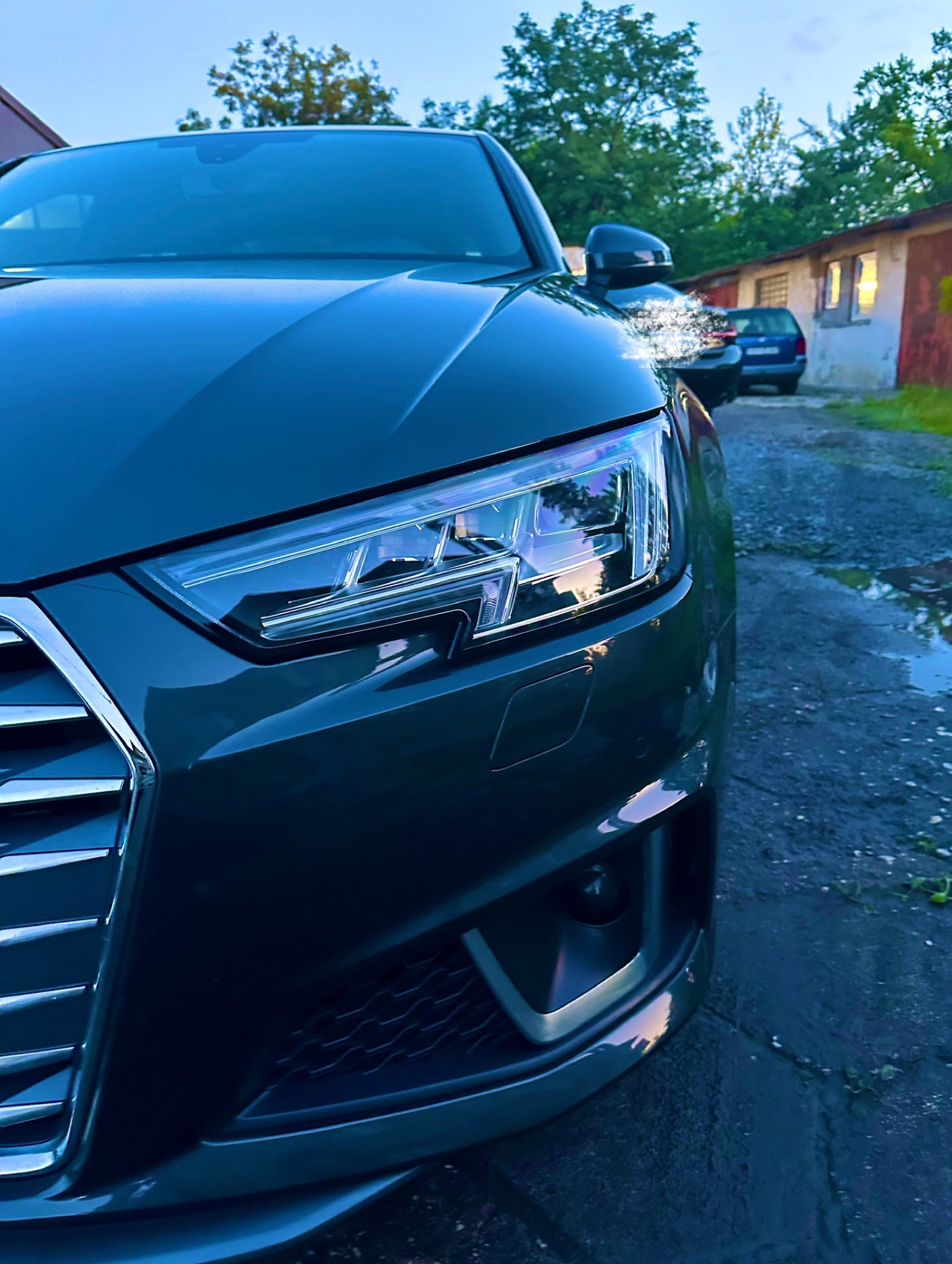 Szary samochód osobowy marki Audi, z reflektorem i grillem, zaparkowany na zewnątrz w pochmurny dzień. Widoczne budynki w tle.