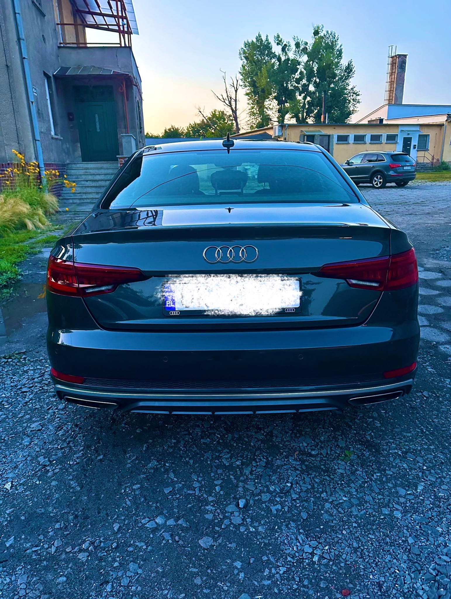 Ciemny sedan Audi A4, widok z tyłu na tle budynku z szarym kamieniem i żwirowego podłoża. Tablica rejestracyjna jest zamazana.