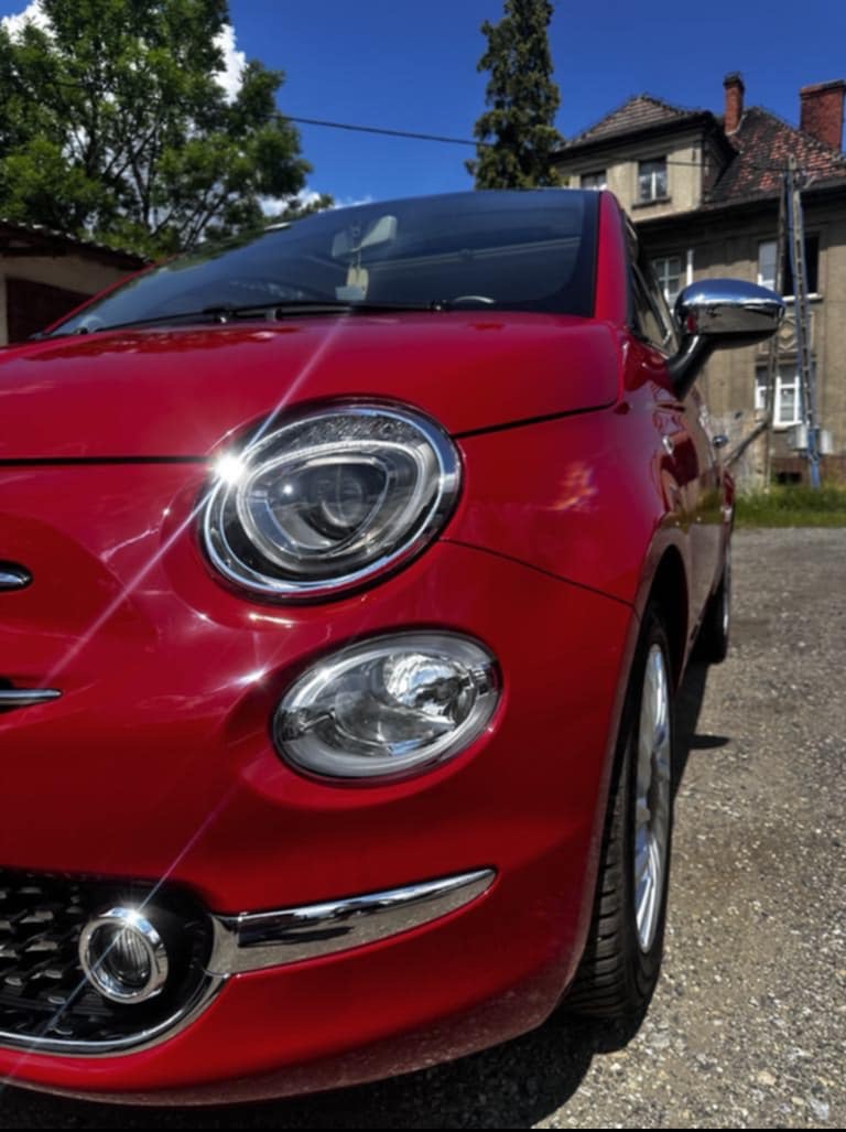 Błyszczący, czerwony Fiat 500 po myciu, z widocznymi refleksami światła na karoserii. W tle budynek z drabiną.