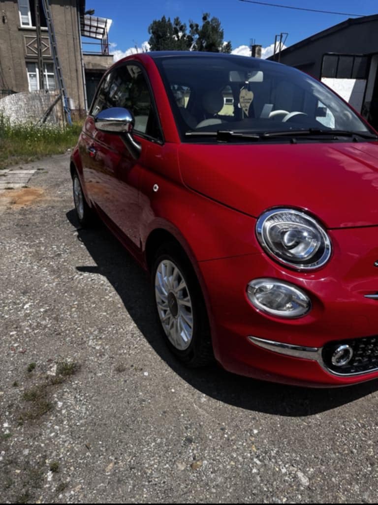 Czerwony Fiat 500 na żwirowym podłożu, uchwycony w słoneczny dzień. Widoczny fragment budynku w tle. Auto z chromowanymi lusterkami i elementami wykończenia.