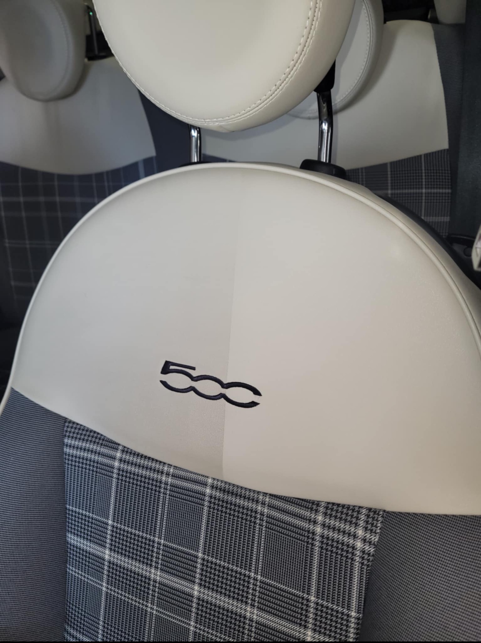 Detal wnętrza auta: białe skórzane siedzenie z logo '500', wykończone szarą kratą. Widoczny zagłówek i fragment pasów bezpieczeństwa. Jasne, równomierne oświetlenie.