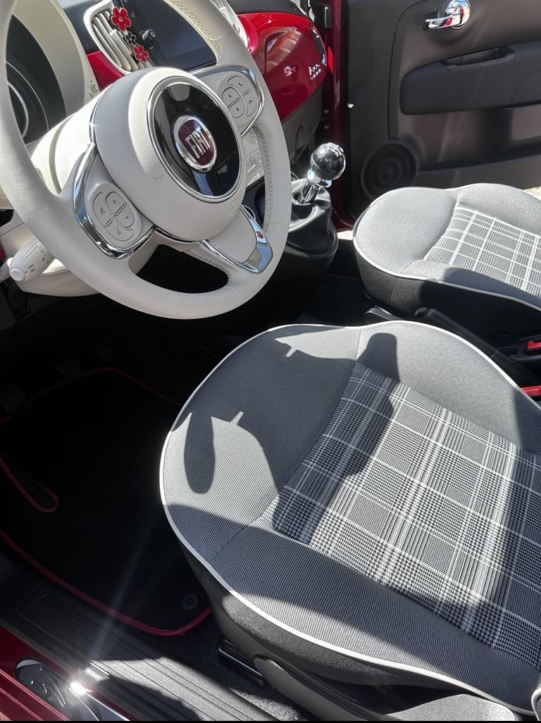 Wnętrze czerwonego Fiata 500 z jasną kierownicą, wzorzystymi fotelami i czarnymi dywanikami z czerwonym obszyciem. Widok na deskę rozdzielczą, dźwignię zmiany biegów i drzwi.