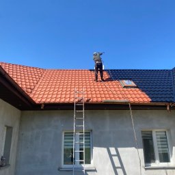 V&A Mal-Bud - Pracownik maluje dach natryskowo na czarno, część dachu pomalowana na czerwono. Na dole drabina oparta o ścianę budynku w surowym stanie.