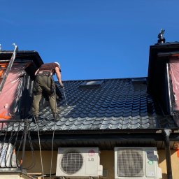 V&A Mal-Bud - Pracownik zabezpieczony linami maluje czarny dach z dachówki ceramicznej. Okna zabezpieczone folią, drabina oparta o dach. Widoczne klimatyzatory na ścianie budynku.