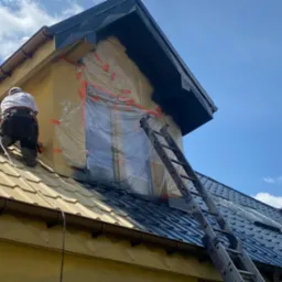 Pracownik maluje dach domu na ciemny kolor. Część dachu pomalowana, okno zabezpieczone folią. Widoczna drabina i fragment elewacji budynku.