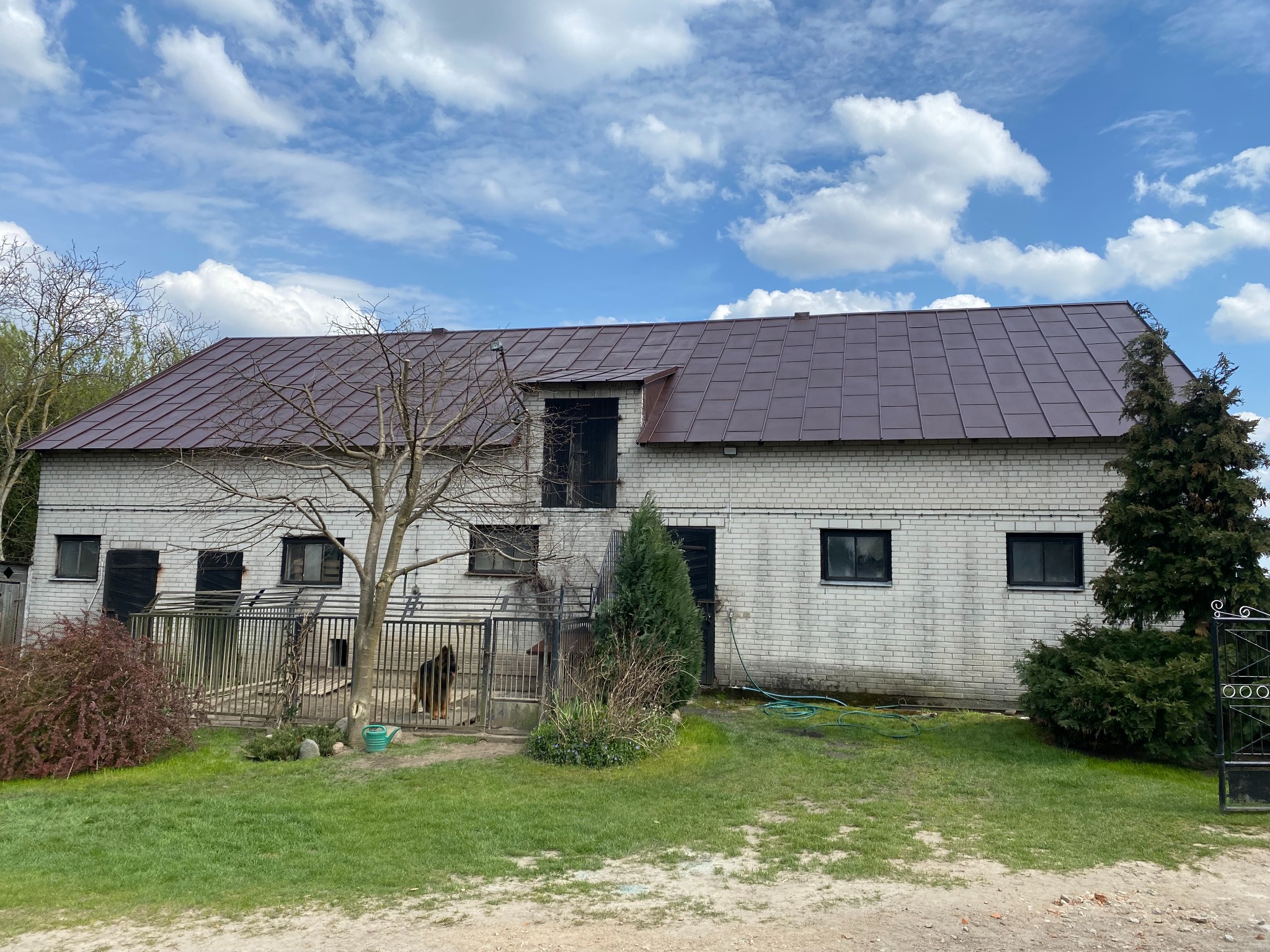 Dwukondygnacyjny budynek z białej cegły z brązowym dachem i czarnymi oknami, otoczony zielenią i drzewami, widziany w słoneczny dzień z lekkim zachmurzeniem.