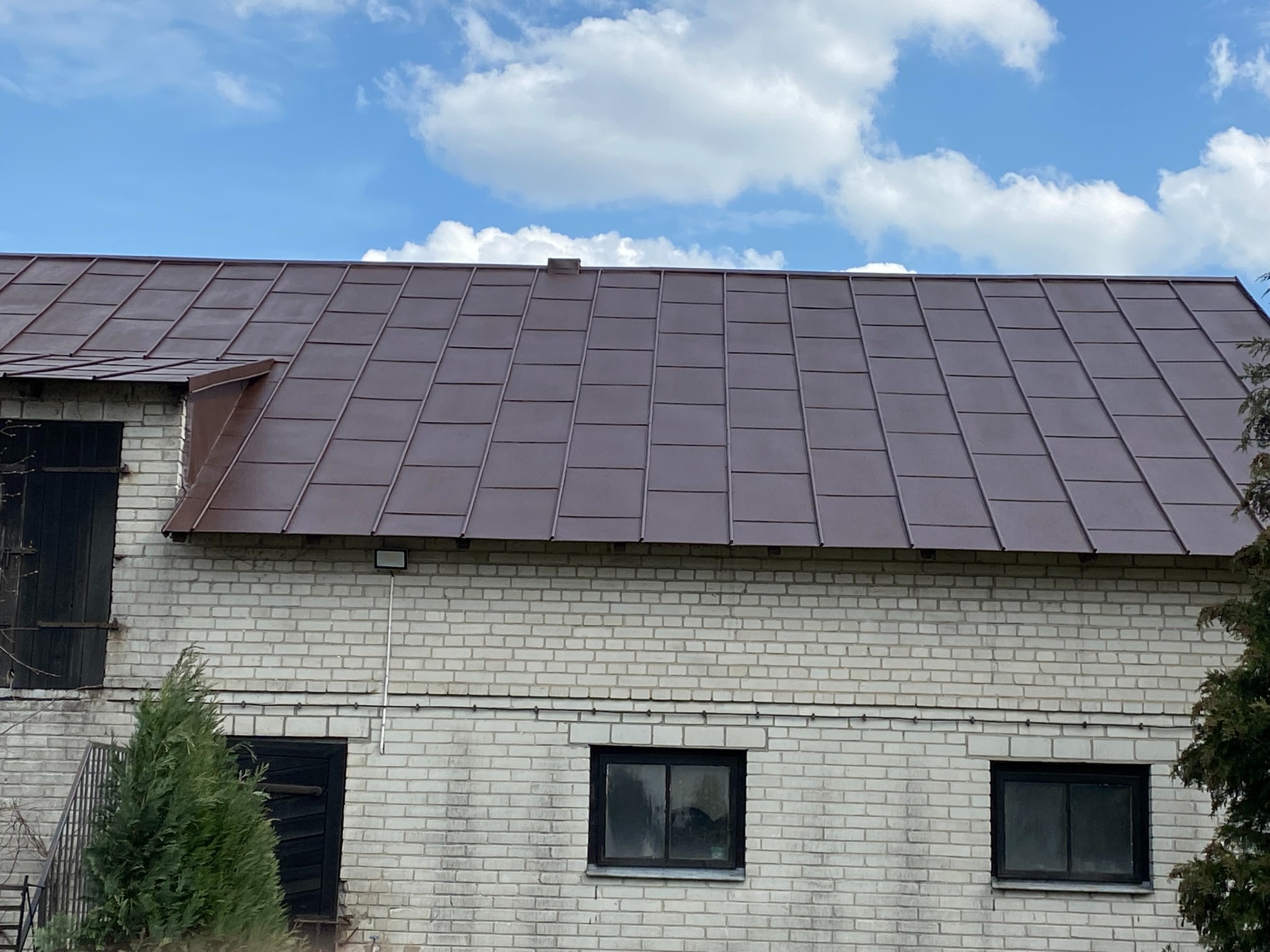 Brązowy, metalowy dach na murowanym budynku z białej cegły pod błękitnym niebem z chmurami. Widoczne okna i drzwi, a także fragmenty zieleni.