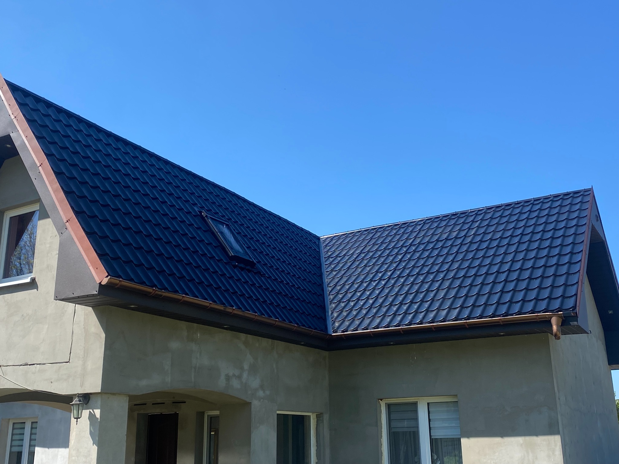 Błyszczący, grafitowy dach z blachodachówki z brązowymi rynnami na surowym budynku w słoneczny dzień. Widoczne okno dachowe i fragment elewacji z oknami.