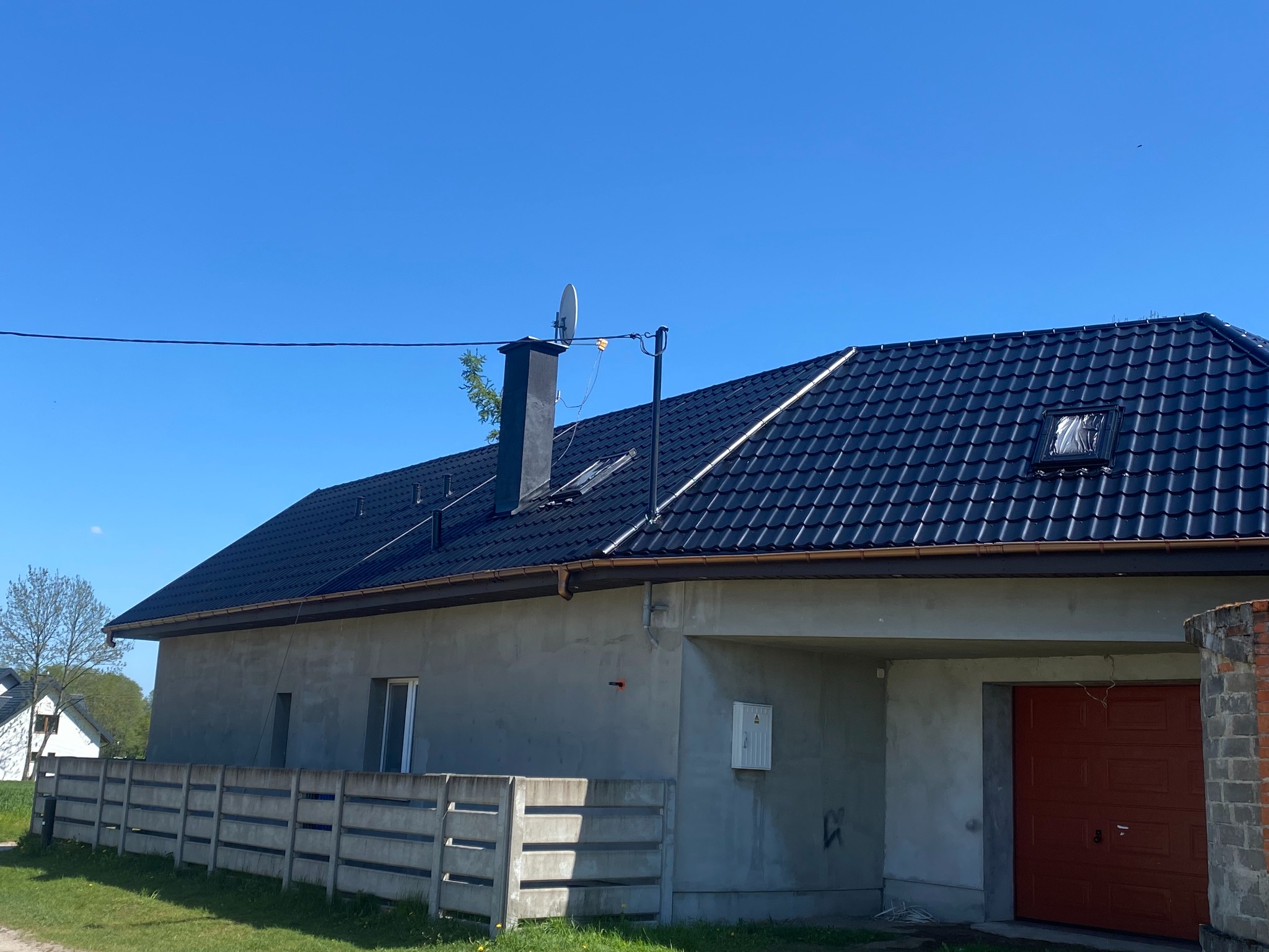 Dom jednorodzinny z ciemnym dachem z dachówki, antena na kominie, Ozorków. Szara elewacja i betonowe ogrodzenie.