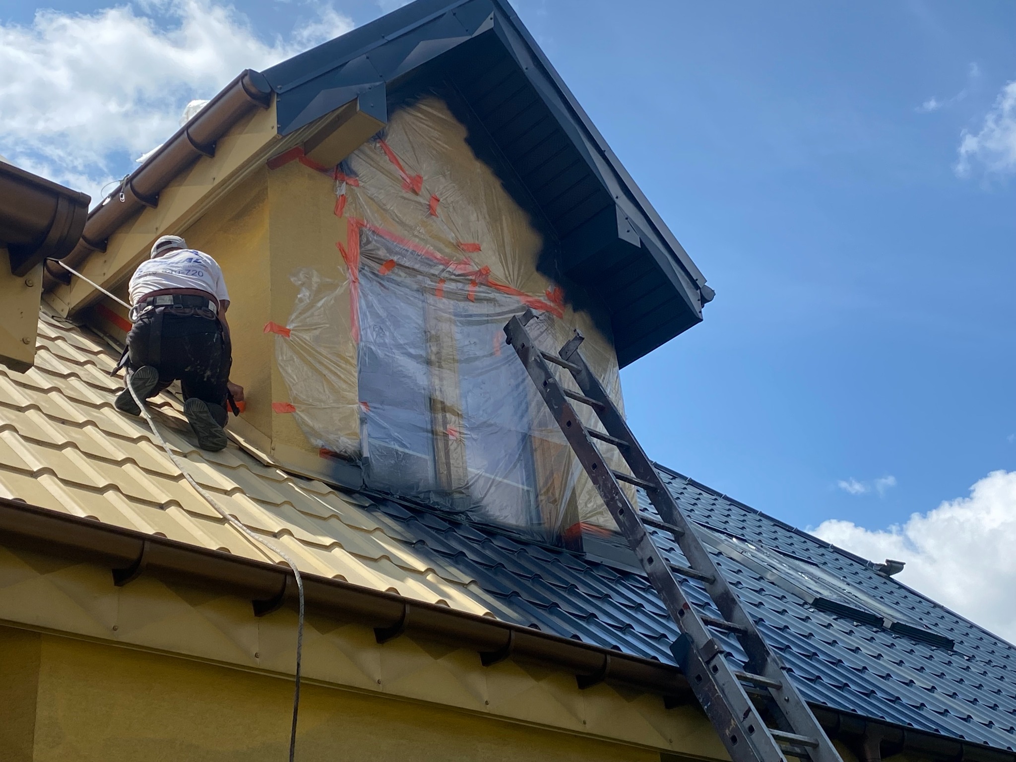 Pracownik maluje dach domu na ciemny kolor. Część dachu pomalowana, okno zabezpieczone folią. Widoczna drabina i fragment elewacji budynku.
