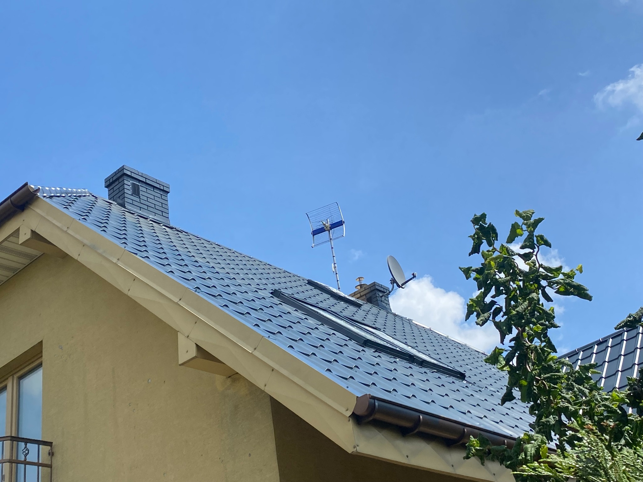Dom z ciemną, błyszczącą dachówką, widok z dołu na dach z kominem, anteną i oknami dachowymi. Jasna elewacja. Błękitne niebo w tle.