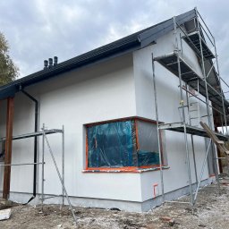 Dmytro Horvat D&Y Vision - Biała elewacja domu jednorodzinnego w trakcie prac wykończeniowych. Widoczne rusztowania, okno zabezpieczone folią i pracownicy podczas malowania.