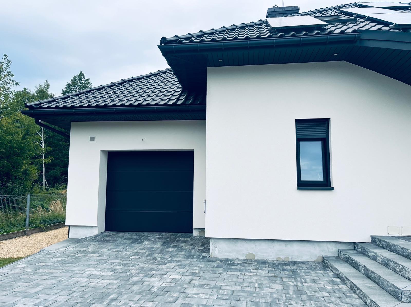 Nowoczesny dom z białą elewacją, czarną bramą garażową i dachem z ciemnej dachówki. Widoczne panele słoneczne i kostka brukowa. Minimalistyczny design.