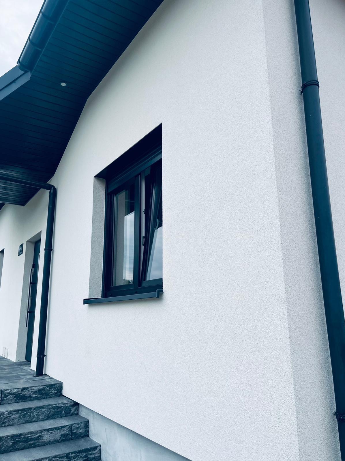 Biała elewacja domu z oknem i rynną w Częstochowie. Nowoczesne wykończenie, widok z dołu, schody wejściowe. Minimalistyczny design.
