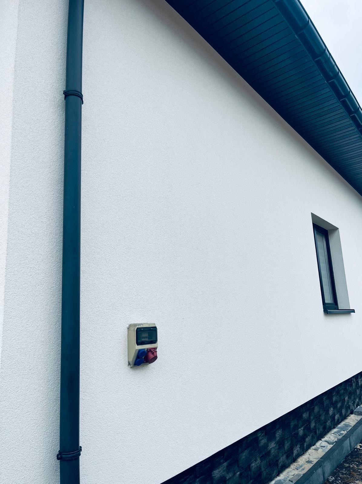 Biała elewacja budynku z ciemnym orynnowaniem i skrzynką elektryczną z gniazdami. Widoczny fragment okna i podmurówki. Tekstura tynku barankowego.