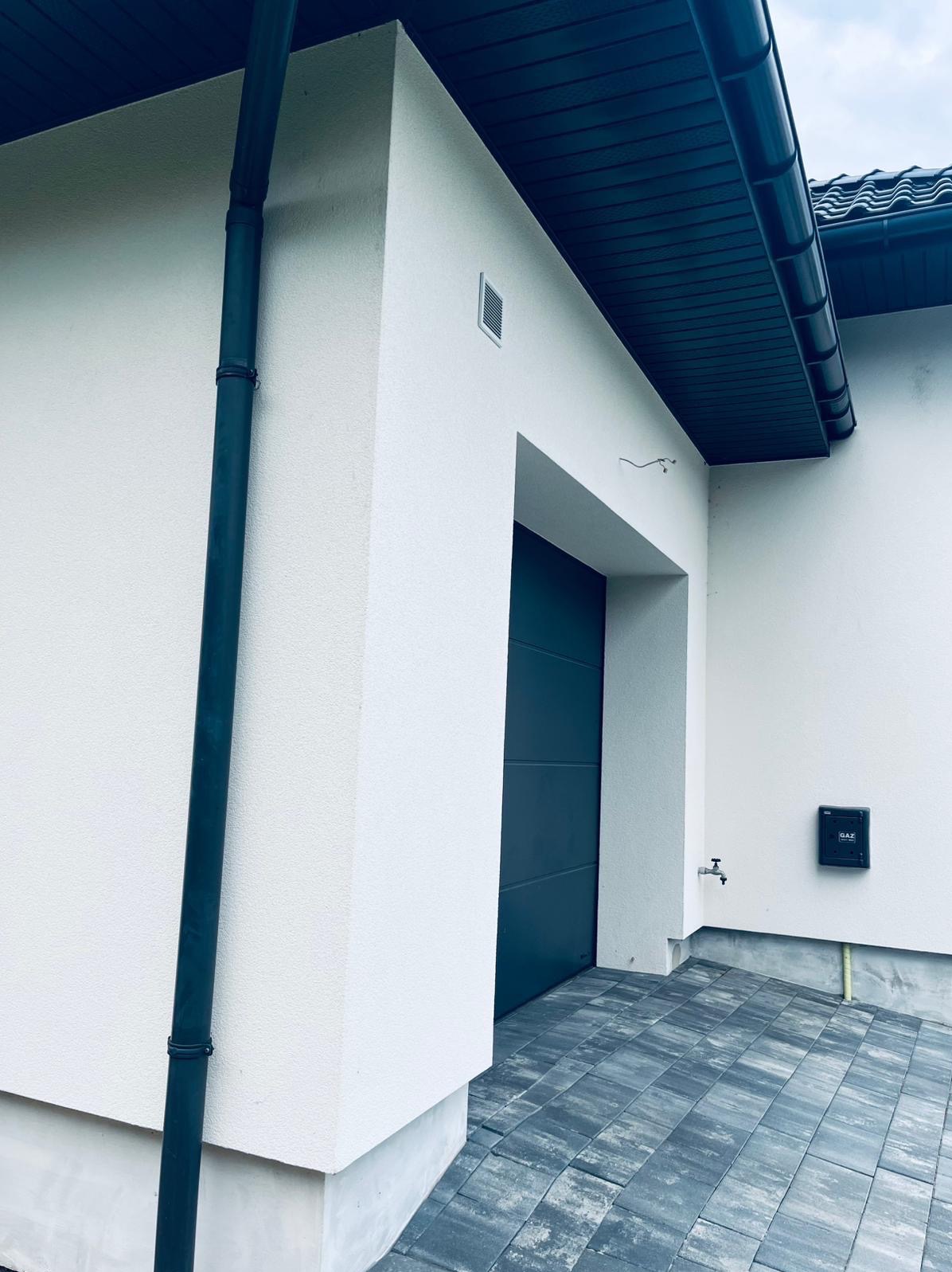 Biała elewacja domu z grafitową bramą garażową i rynną. Kostka brukowa w odcieniach szarości. Skrzynka gazowa na ścianie. Minimalistyczny design.