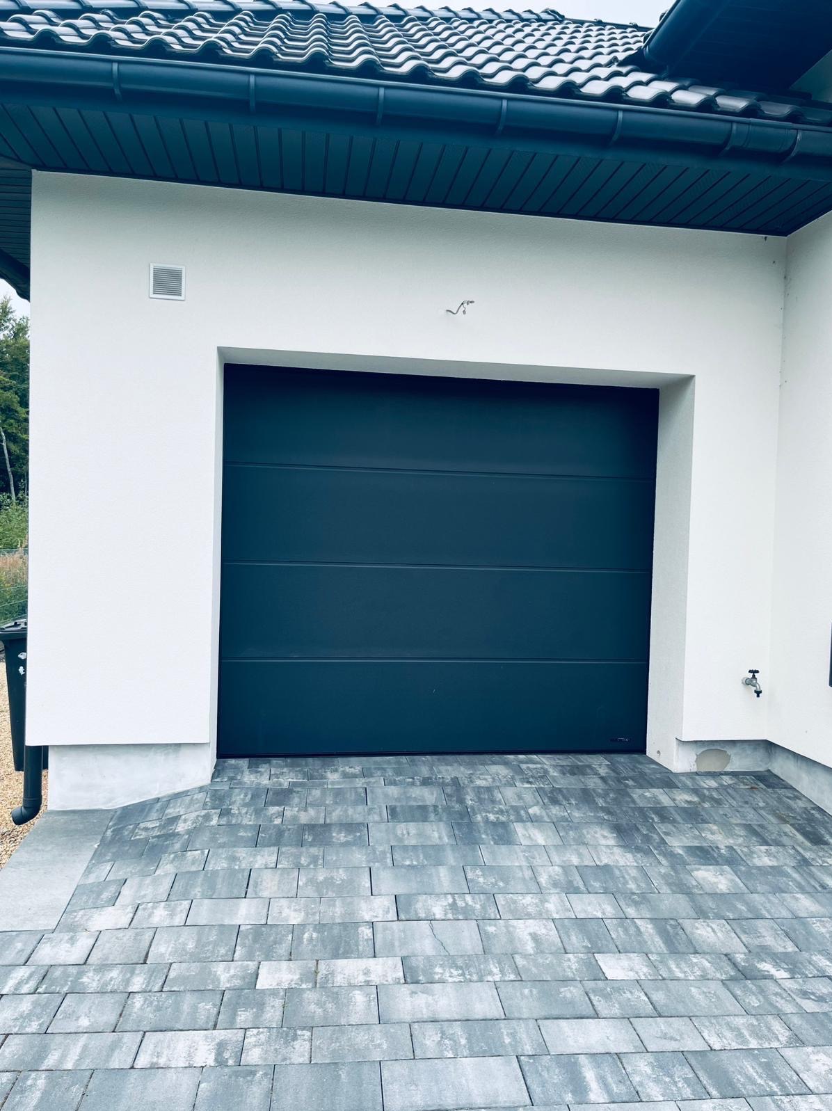 Nowoczesna, grafitowa brama garażowa segmentowa w białej elewacji budynku z kostką brukową. Widoczny fragment dachu z czarną dachówką.