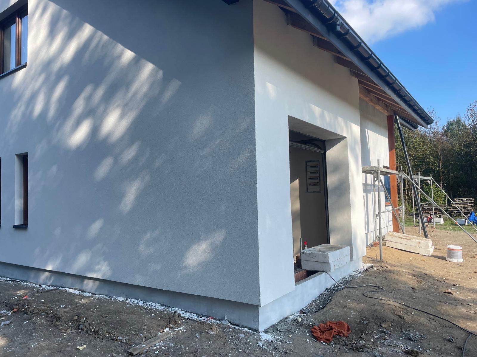 Świeżo otynkowana, jasnoszara elewacja domu jednorodzinnego z widocznym wejściem do garażu i rusztowaniem w tle, w słoneczny dzień. Detal wykończenia.