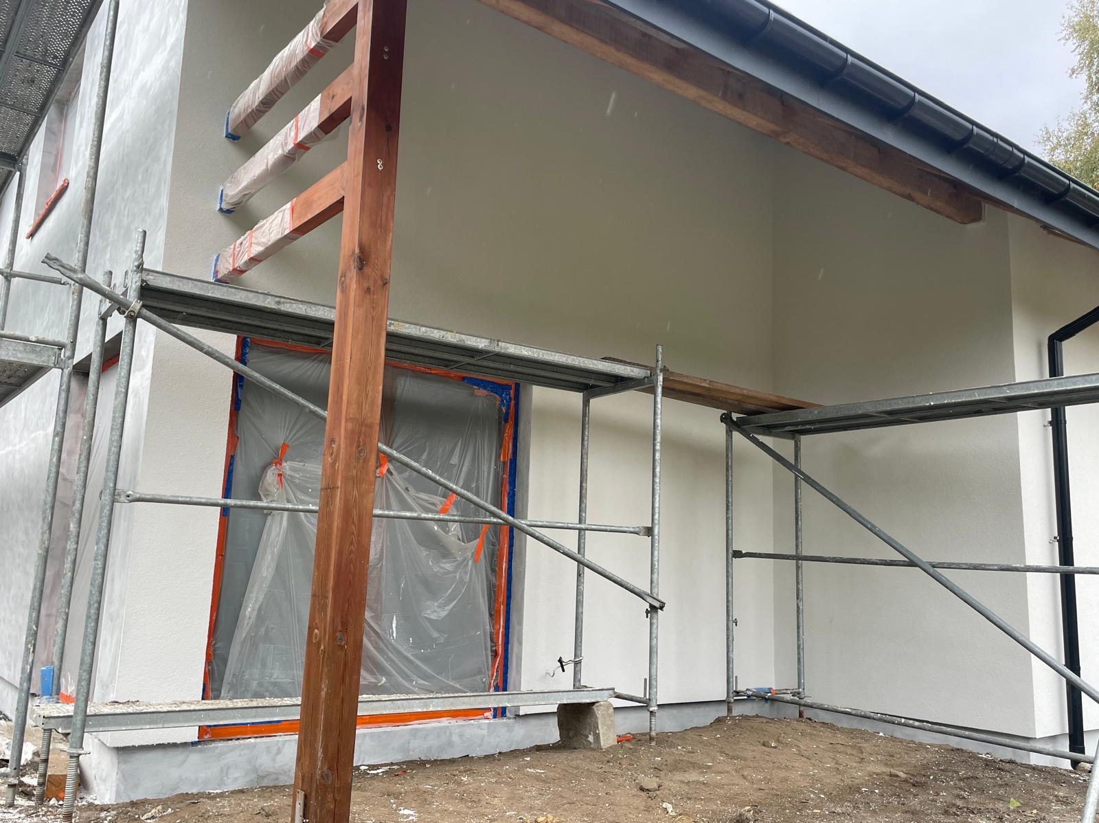 Świeżo otynkowana, biała elewacja domu w trakcie prac wykończeniowych. Okno zabezpieczone folią, widoczne rusztowania i drewniane elementy konstrukcyjne.