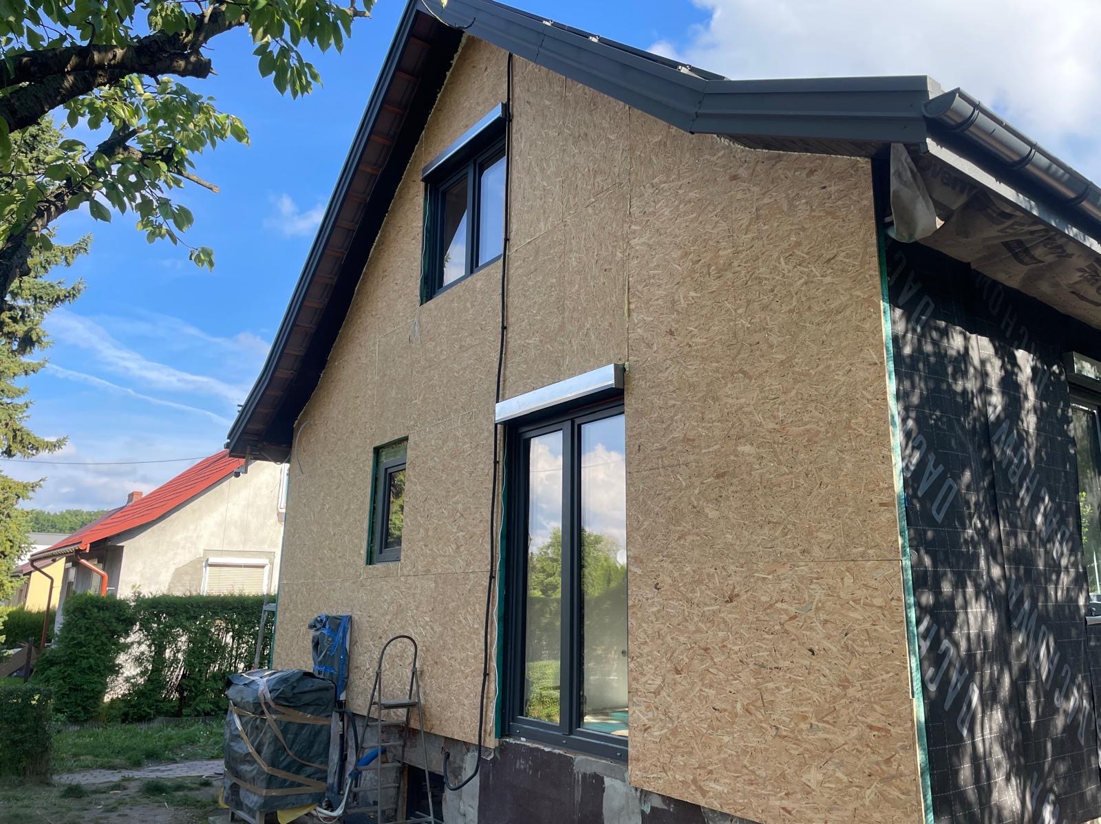 Dom w trakcie ocieplania: płyty OSB na elewacji, okna z roletami zewnętrznymi. Widoczne rusztowanie i materiały budowlane. Częściowo ocieplona ściana z membraną.