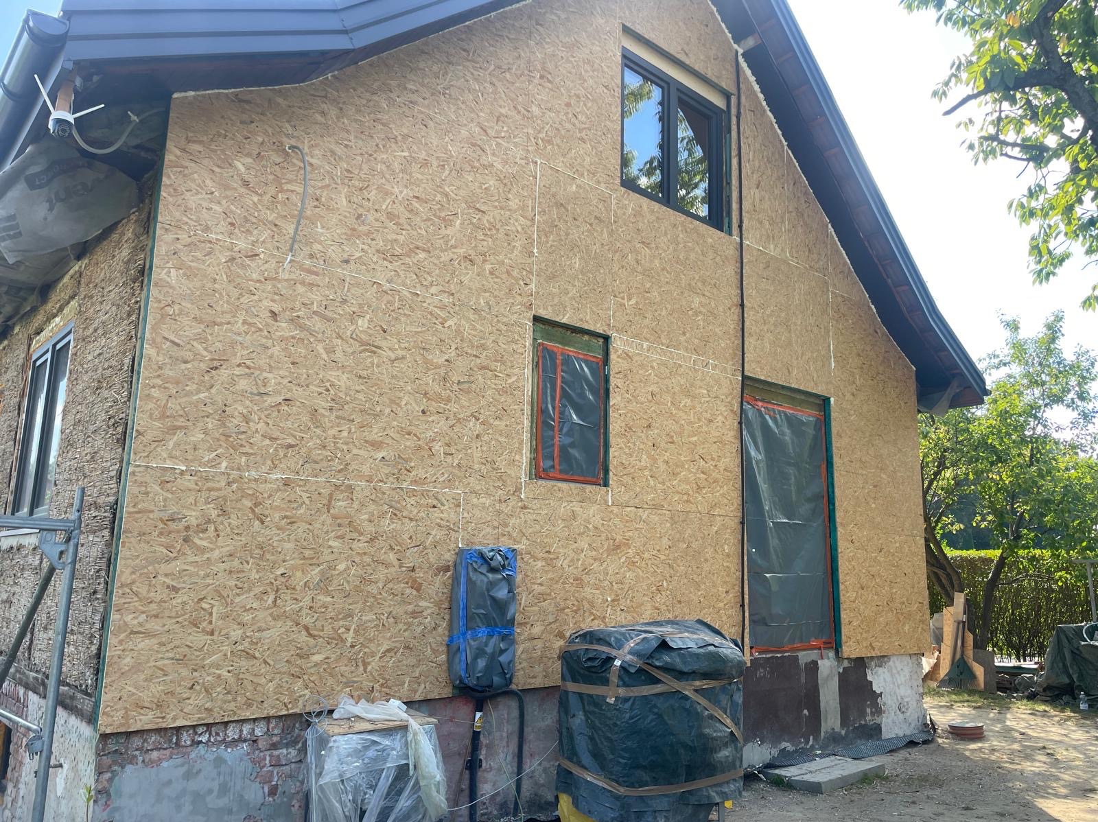 Dom w trakcie ocieplania: płyty OSB na elewacji, okna zabezpieczone folią. Widoczne materiały budowlane i fragment rusztowania. Prace przygotowawcze do dalszych etapów.