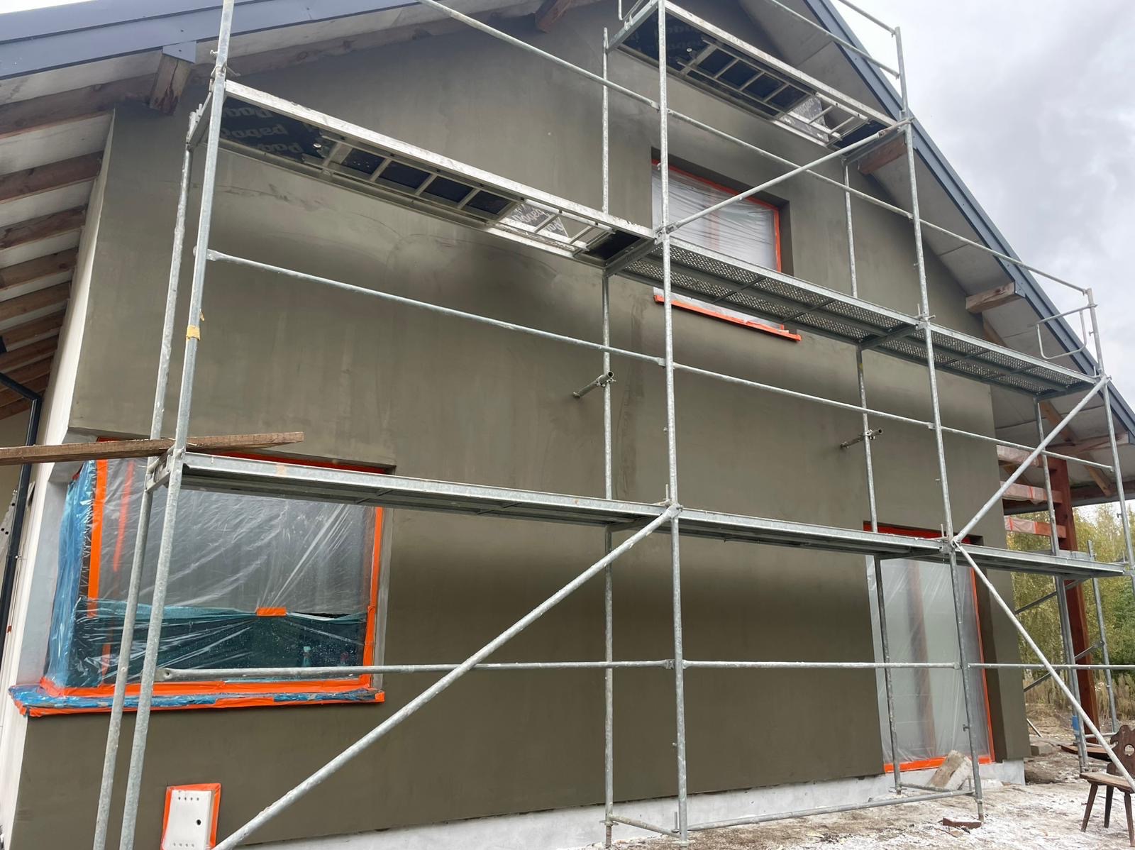 Dom w trakcie prac elewacyjnych: nałożony tynk, okna zabezpieczone folią, rusztowanie. Widok z boku, pochmurne niebo w tle. Etap przed nałożeniem farby.