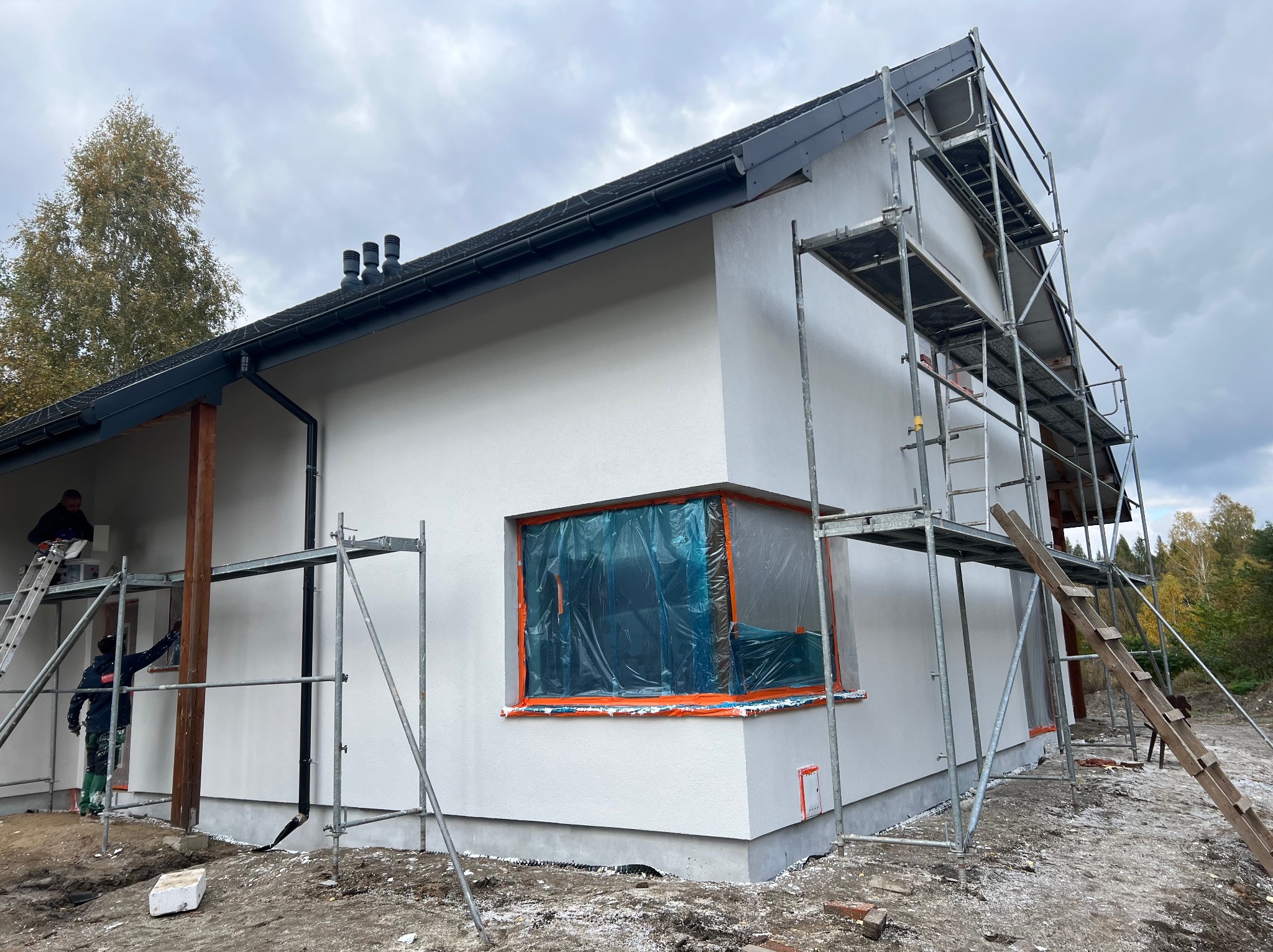 Biała elewacja domu jednorodzinnego w trakcie prac wykończeniowych. Widoczne rusztowania, okno zabezpieczone folią i pracownicy podczas malowania.