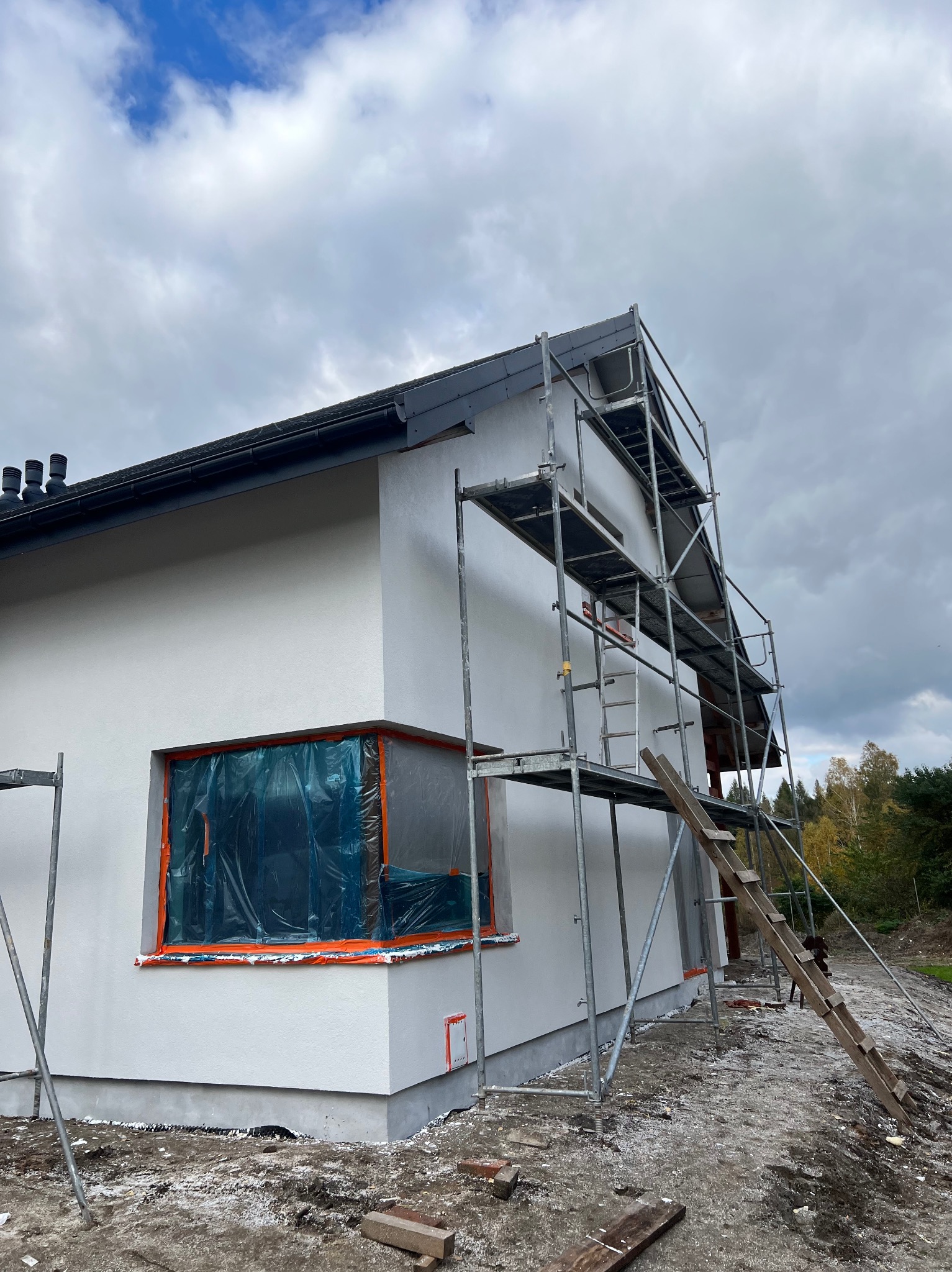 Dom jednorodzinny z nową, białą elewacją, zabezpieczonym oknem folią i rusztowaniem. Widoczne świeże tynki i przygotowania do dalszych prac wykończeniowych.