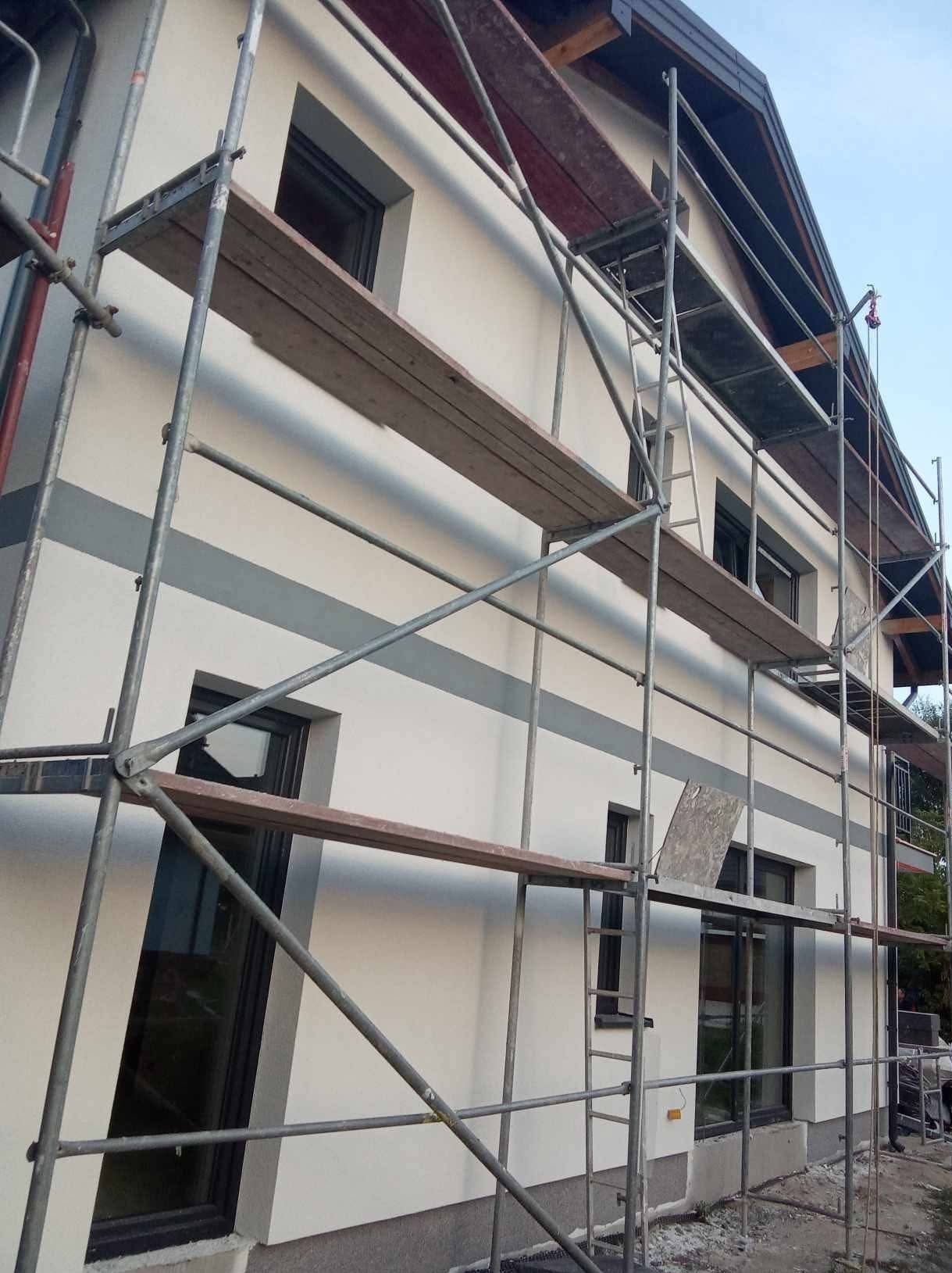 Świeżo otynkowana elewacja domu jednorodzinnego z szarymi pasami, widoczna spod rusztowania. Okna z ciemnymi ramami. Częściowo widoczny dach.