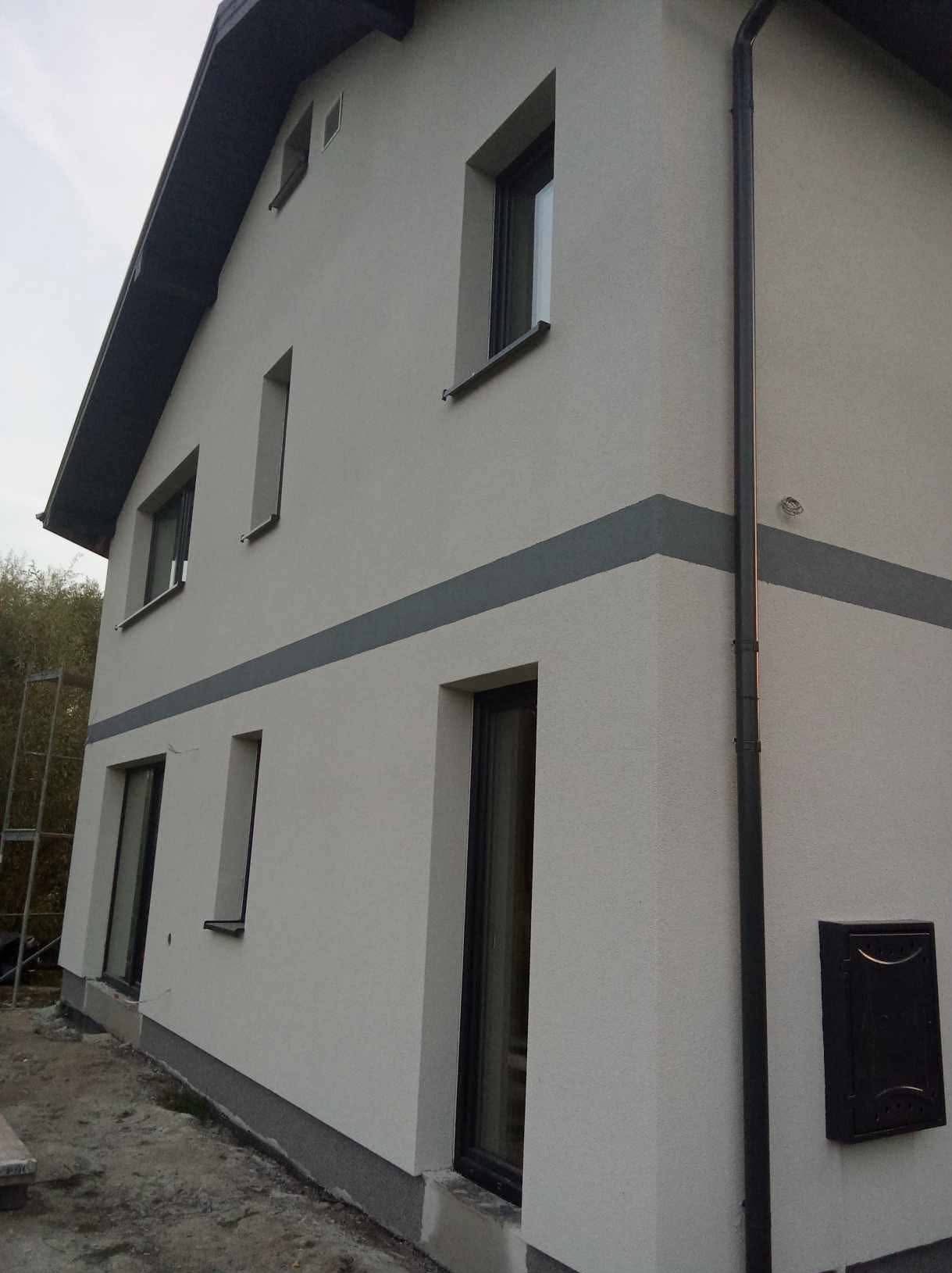 Jasna elewacja domu jednorodzinnego z szarym pasem dekoracyjnym, widok z dołu, rynna pionowa, skrzynka na listy, okna z czarnymi ramami.