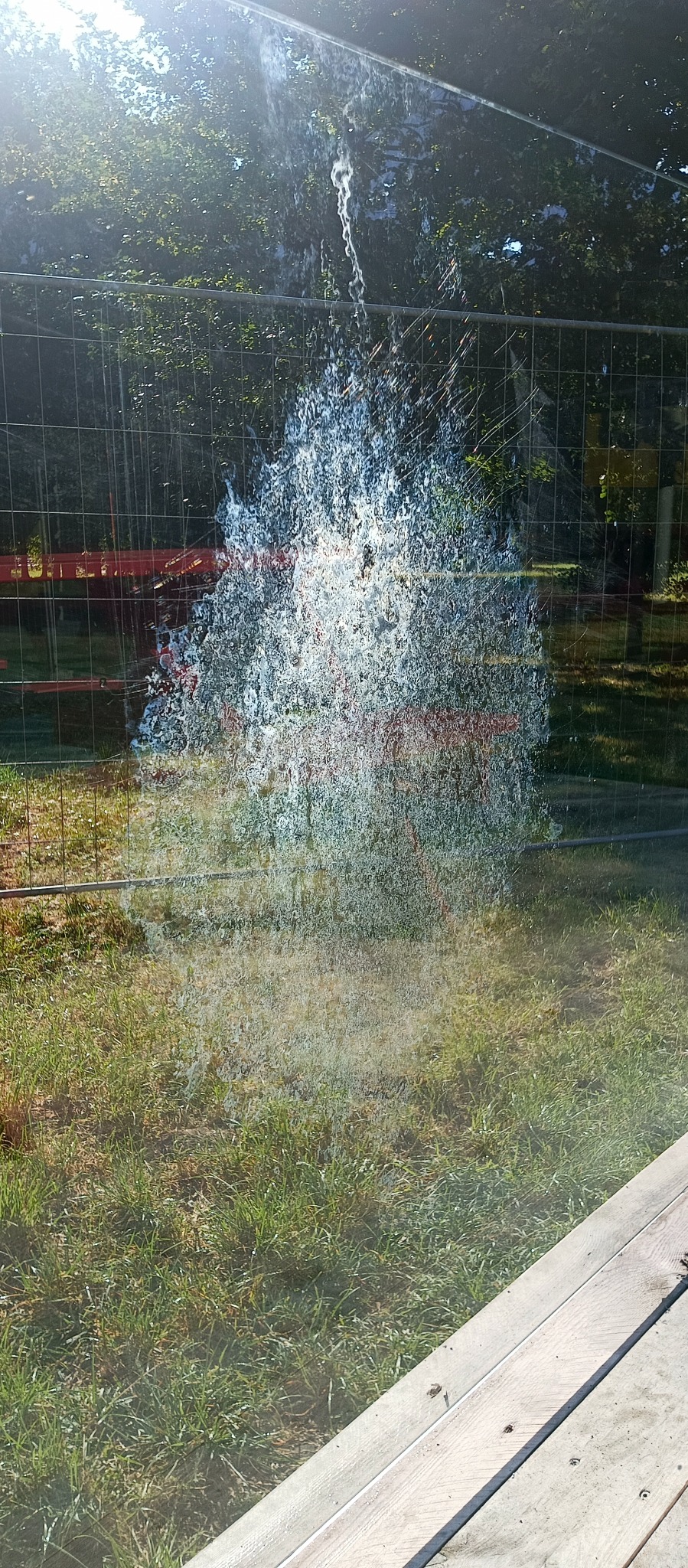 Zabrudzone okno z widocznymi smugami po deszczu lub czyszczeniu, na tle zieleni i ogrodzenia budowlanego. Widoczne odbicia światła.