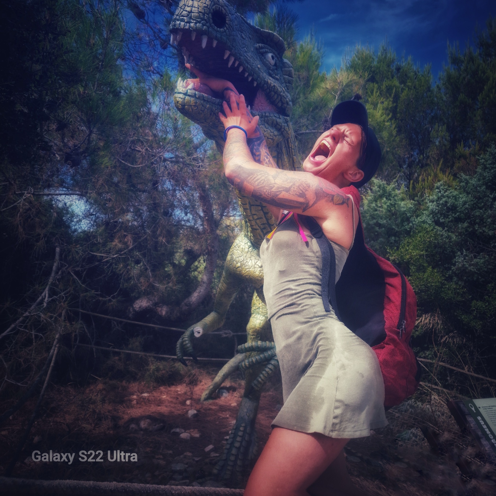 Kobieta z tatuażami udaje atak na rzeźbę dinozaura w parku, szeroko otwarte usta, w tle drzewa i niebo. Nietypowa sesja zdjęciowa w plenerze.