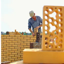 Murarz układa ścianę z dziurawki, na pierwszym planie cegła z otworami. Praca na zewnątrz, w tle widać zieleń i niebo.