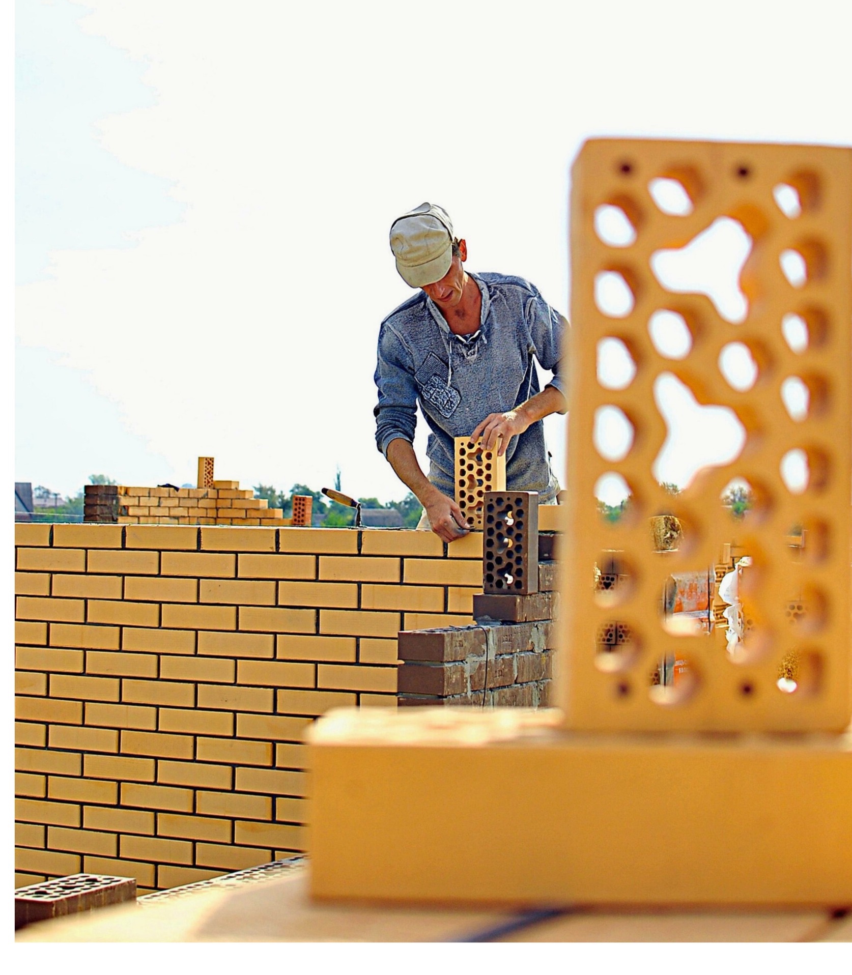 Murarz układa ścianę z dziurawki, na pierwszym planie cegła z otworami. Praca na zewnątrz, w tle widać zieleń i niebo.