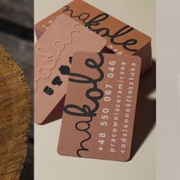 Ilona Mrozowska Grafik Komputerowy - Brązowe wizytówki z tłoczonym logo 'nakole' i danymi kontaktowymi pracowni ceramicznej, ułożone obok drewnianego elementu i etykiety z fragmentem tekstu.